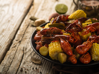 Rough wooden table. On it we see a large black bowl with pieces of boiled corn and crayfish. In the background is half a lemon. Close-up. High angle view.