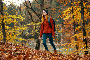 happy woman hiker with a backpack on her back in jeans and a red sweater in the autumn forest park landscape