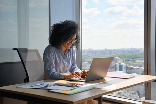 Happy Smiling African American Businesswoman Executive Manager Sitting At Desk Working Typing On Laptop Computer In Contemporary Corporation Office With Panoramic View. Business Technologies Concept.