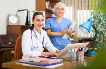 Young woman doctor and nurse working together with case histories on laptop in modern office