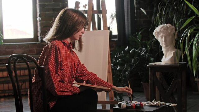 A young pretty woman artist sits in front of an easel and mixing warm colors on a wooden palette