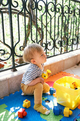 Little girl sits on a rug on the balcony and holds toys in her hand