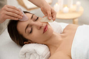 Fototapeta na wymiar Young woman receiving facial massage with gua sha tools in beauty salon