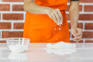 Unrecognizable  woman in orange apron smile and bend to enjoy kneading dough by hands beside glass bowl of flour powder on white cooking table as food preparation near brick wall in home kitchen