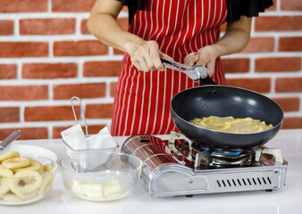 Woman chef in red striped apron cooking near brick wall by holding metal tongs and black pan on hot gas stove for frying potatoes on table with some glass bowl of ingredients in home kitchen