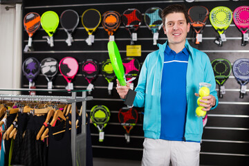 Sporty adult male in uniform is holding new rocket and balls for padel in the store.