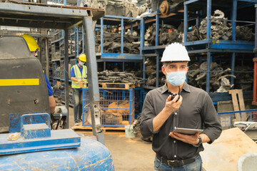 Men work together, wear safety facemask, hold tablet, using walkie-talkie. Caucasian engineer men hold tablet, using walkie-talkie while Asian men working behind him in factory-warehouse