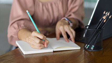 Close up view of female hand writing on notebook while working with digital tablet