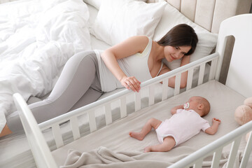 Happy young mother near crib with sleeping newborn baby in bedroom - obrazy, fototapety, plakaty