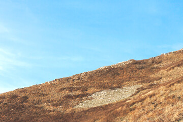 View of a hill covered with yellow and brown dry grass and stones against a blue sky with white clouds