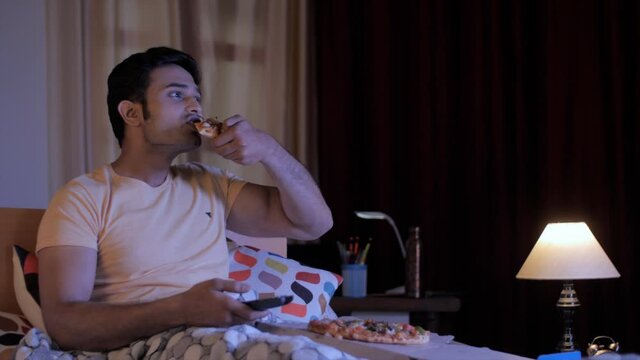 Portrait of a young man sitting and relaxing on the bed watching television - Binge eating . Medium shot of a hungry Indian male eating a slice of pizza while watching TV at night