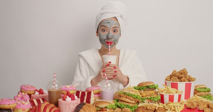 Young female model applies clay mask on face for healthy skin drinks soda looks surprisingly at great variety of junk food dressed in bathrobe has unhealthy eating habits isolated on white background