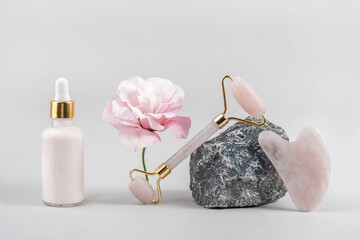 Crystal rose quartz facial roller, cosmetic dropper bottle on stone and beautiful flower on grey...