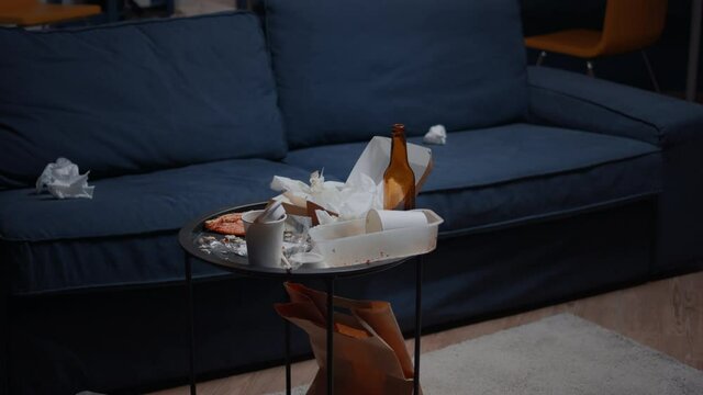 Empty home of depressed person with messy table with leftover thrown on floor in unorganized living room of depresive person. Pizza, beer bottles and napkins in dirty apartment
