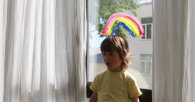 Preschooler boy in yellow t-shirt stands near window with painted rainbow at bright sunlight between white curtains at home