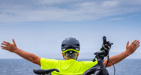 Rear view of senior cyclist man resting at sea on the cliff after excursion with his electro bike....