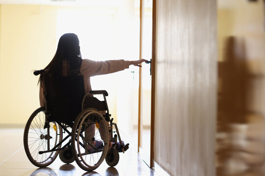Woman in wheelchair reaches out to open handle of door in dark hallway - Powered by Adobe