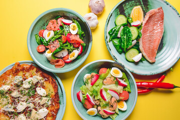 Fototapeta na wymiar Salad with spinach, eggs and radish in a bowl over bright yellow background. Fish fillet steak, potato and cheese pizza.