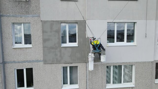 An industrial climber suspended on ropes and paints the wall of a building with a roller. A male worker performs work on the insulation of the facade of a multi-storey building. View from the drone.