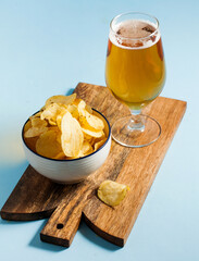 Chilled light beer in a glass and natural chips in a bowl on a wooden board on a blue background. 