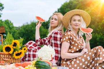 Friends is making picnic outdoor. Laughing girls sitting on white knit picnic blanket drinking wine. Happy young women talking, smiling outdoors on Summer picnic on sunny day at park 