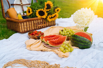 Summer picnic concept on sunny day with watermelon, fruit, bouquet hydrangea and sunflowers flowers. Picnic basket on grass with food and refreshing summer drink on white knit blanket. Selective focus