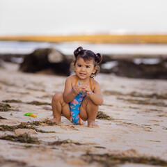 Cute little girl sitting on sandy beach, playing with sand. Happy childhood. Summer vacation. Holiday concept. Baby girl wearing blue swimsuit. Bali, Indonesia