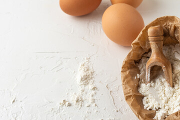 Aerial view of paper bag with flour and wooden spoon on white wooden table with brown eggs, horizontal, with copy space