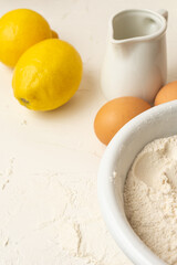 Top view of bowl with flour, eggs, white jug and lemons, on white table, selective focus, vertical, with copy space