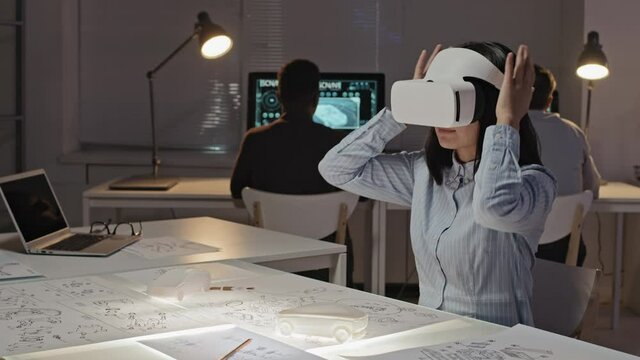 Slowmo shot of female Asian engineer putting VR headset on and gesticulating while working on project in virtual reality D printed car models and blueprints are on table