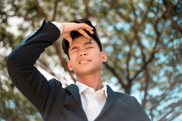 A young absentminded asian university student clenches his forehead in frustration, possibly forgetting something or failed a subject. At the park or campus grounds. - obrazy, fototapety, plakaty