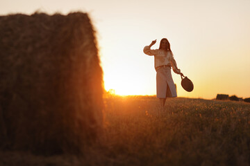 young woman in the beautiful light of the summer sunset in a field is walking near the straw bales. beautiful romantic girl with long hair outdoors in field.