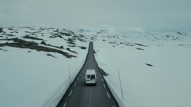 Drone aerial shot, camera follows cute vintage camping van driving through snowy mountain winter road. Adventure camping RV vanlife lifestyle in scandinavian countries