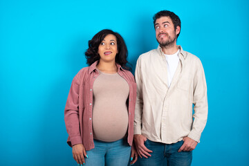 Amazed young couple expecting a baby standing against blue background bitting lip and looking...
