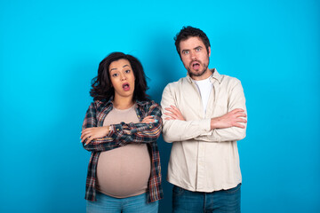 Shocked embarrassed young couple expecting a baby standing against blue background keeps mouth widely opened. Hears unbelievable novelty stares in stupor