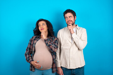 Naklejka premium Thoughtful young couple expecting a baby standing against blue background holds chin and looks away pensively makes up great plan