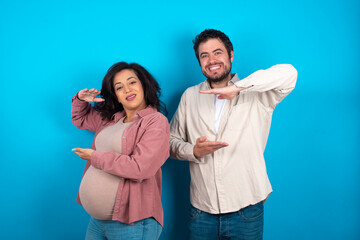 young couple expecting a baby standing against blue background gesturing with hands showing big and large size sign, measure symbol. Smiling looking at the camera.