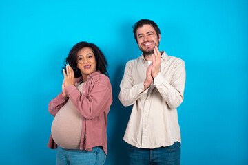 young couple expecting a baby standing against blue background clapping and applauding happy and...