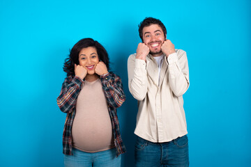 Happy young couple expecting a baby standing against blue background  keeps fists on cheeks smiles broadly and has positive expression being in good mood