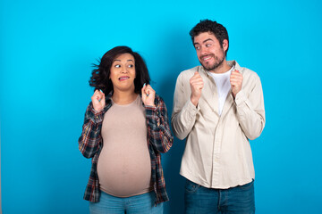 young couple expecting a baby standing against blue background clenches fists and awaits for something nice happened looks away bites lips and waits announcement of results