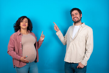 Smiling young couple expecting a baby standing against blue background indicating finger empty space showing best low prices