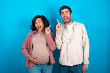 young couple expecting a baby standing against blue background holding finger up having idea and posing