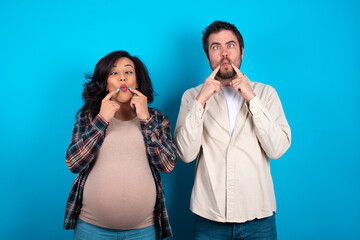young couple expecting a baby standing against blue background crosses eyes and makes fish lips...