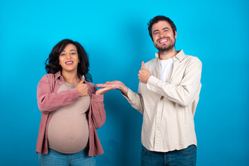 young couple expecting a baby standing against blue background Showing palm hand and doing ok gesture with thumbs up, smiling happy and cheerful.