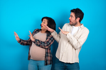 Displeased young couple expecting a baby standing against blue background keeps hands towards empty space and asks not come closer sees something unpleasant