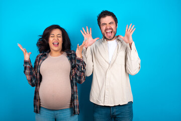 Crazy outraged young couple expecting a baby standing against blue background screams loudly and gestures angrily yells furiously. Negative human emotions feelings concept