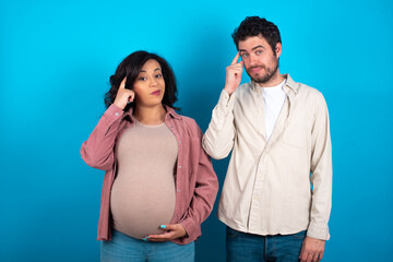 Naklejka premium young couple expecting a baby standing against blue background tries to memorize something, keeps fore finger on temple, reminds information for exam,