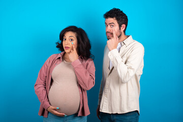 Naklejka premium young couple expecting a baby standing against blue background Pointing to the eye watching you gesture, suspicious expression.