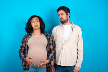 Dissatisfied young couple expecting a baby standing against blue background purses lips and has unhappy expression looks away stands offended. Depressed frustrated model.