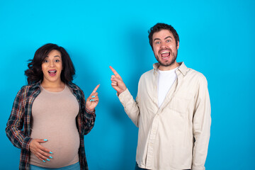 young couple expecting a baby standing against blue background points aside on copy blank space. People promotion and advertising concept